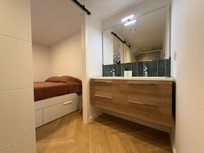 A bedroom with a bed, a wooden dresser, a mirror, and a bathroom with a sink and mirror. - PEONTENIA - BIDARRAY (BIDARRAY)