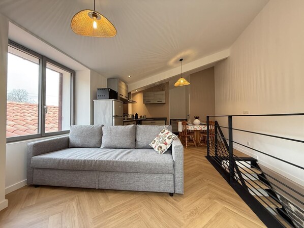 A modern living room with a grey sofa, a dining area, and a kitchen in the background. - PEONTENIA - BIDARRAY (BIDARRAY)