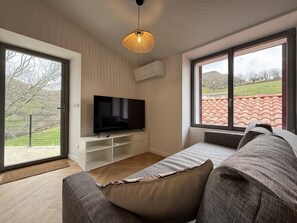 A modern living room with a flat-screen TV, a sofa, and a large window offering a view of a red-tiled roof and greenery. - PEONTENIA - BIDARRAY (BIDARRAY)