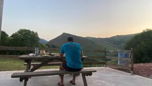 A person seated at a wooden table with a view of a hilly landscape. - PEONTENIA - BIDARRAY (BIDARRAY)