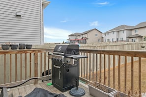 A deck with a barbecue grill, potted plants, and a view of residential buildings.