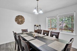 Comedor con mesa de madera oscura, seis sillas, candelabro, un adorno en la pared y ventana con vista a follaje.