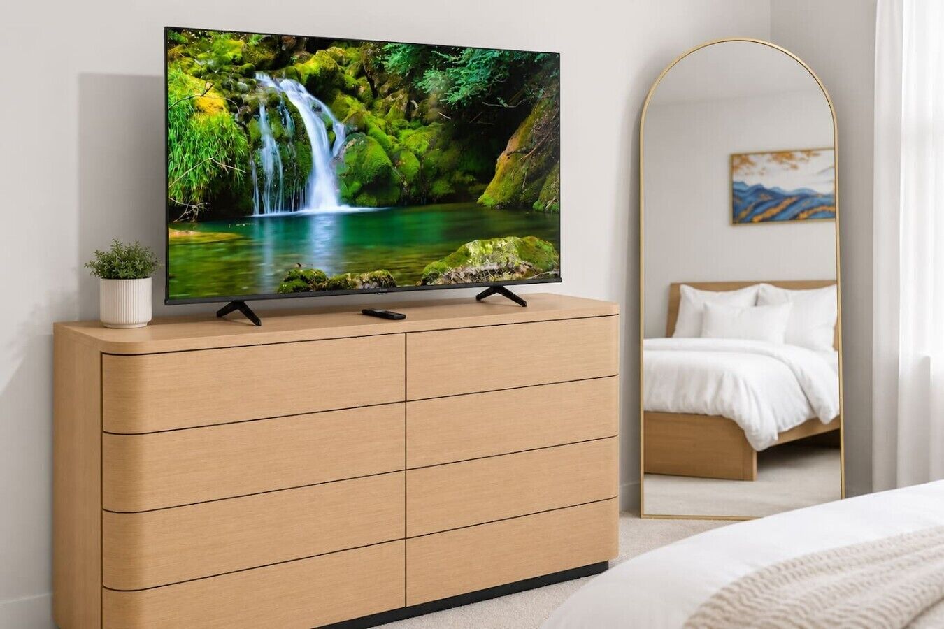 A TV on a wooden dresser with a remote, a potted plant, and a bedroom with a bed and a painting in the background.