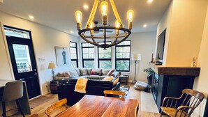 A dining area with a wooden table, chairs, a chandelier, and a fireplace. - Le Refuge Douillet in the heart of Tremblant: ski, bike, pool and spa (Mont-Tremblant)