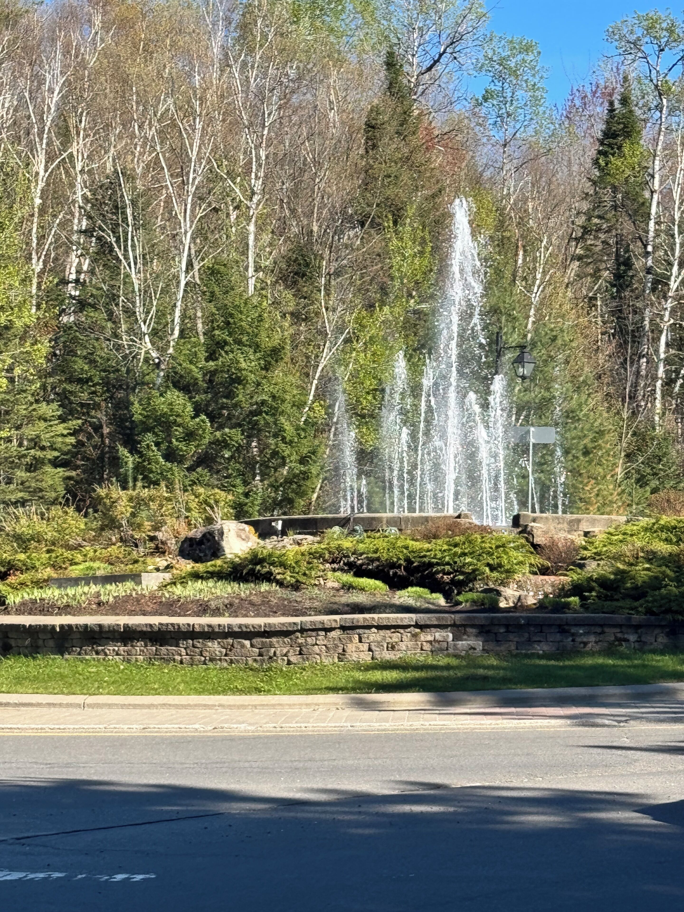 Una fuente con chorros de agua en un área ajardinada con árboles y un muro de piedra.