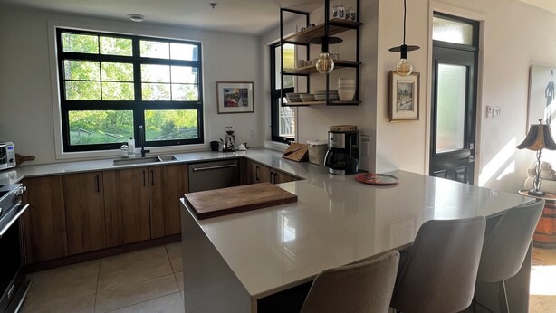 A modern kitchen with a large island, bar stools, a built-in oven, and a window overlooking greenery. - Le Refuge Douillet in the heart of Tremblant: ski, bike, pool and spa (Mont-Tremblant)