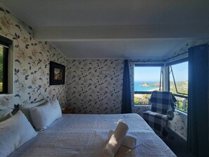 A bedroom with a bed, pillows, a chair, a window with a view, and a framed picture on the wall.