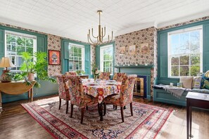Ein Esszimmer mit Tisch, gedeckt für sechs Personen, umgeben von stuhlartigen Sitzgelegenheiten mit Blumenmuster, einer Kristallleuchter und einem Kamin.