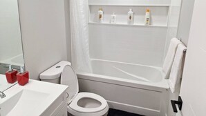Un baño con inodoro blanco, un lavabo con dispensadores de jabón rojo, una ducha con cortina blanca y un estante con artículos de aseo.