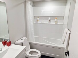 A bathroom with a white toilet, a sink with red soap dispensers, a shower with a white curtain, and a shelf with toiletries. - Cozy Two Bedrooms Basement Suite (Winnipeg)