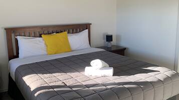 Una cama bien tendida con colcha gris, cabecero de madera, almohadas blancas y amarillas, y una toalla enrollada sobre la cama.