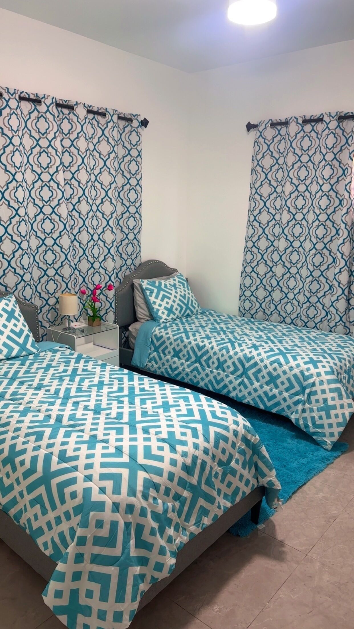 Une chambre avec deux lits, des rideaux à motifs, une table de chevet avec une lampe et une petite plante en pot.