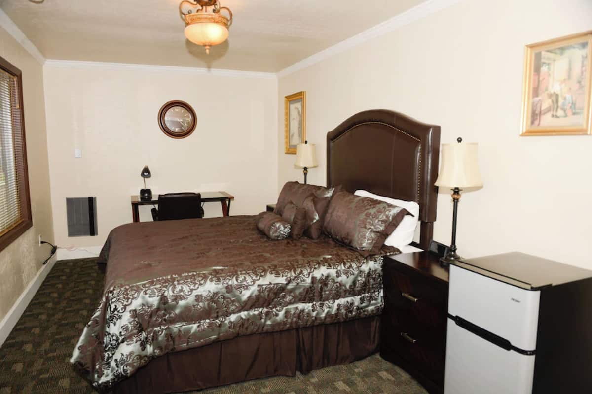 A bedroom with a bed, bedside table, lamp, clock, and a small refrigerator.