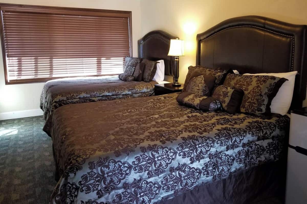 A hotel room with two beds, a nightstand, a lamp, and a window with blinds.