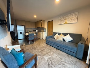 Ein modernes Wohnzimmer mit grauem Sofa, einem Essbereich mit Tisch und Stühlen, einer Küche mit Mikrowelle und Herd sowie einem dekorativen Baum-Kunstwerk an der Wand.