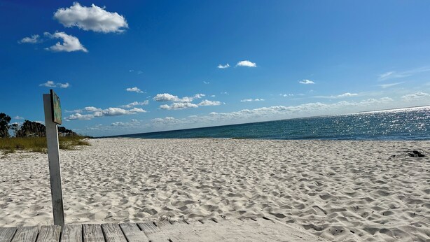 Ein Sandstrand mit einer hölzernen Prominade, die zum Wasser führt, unter blauem Himmel.