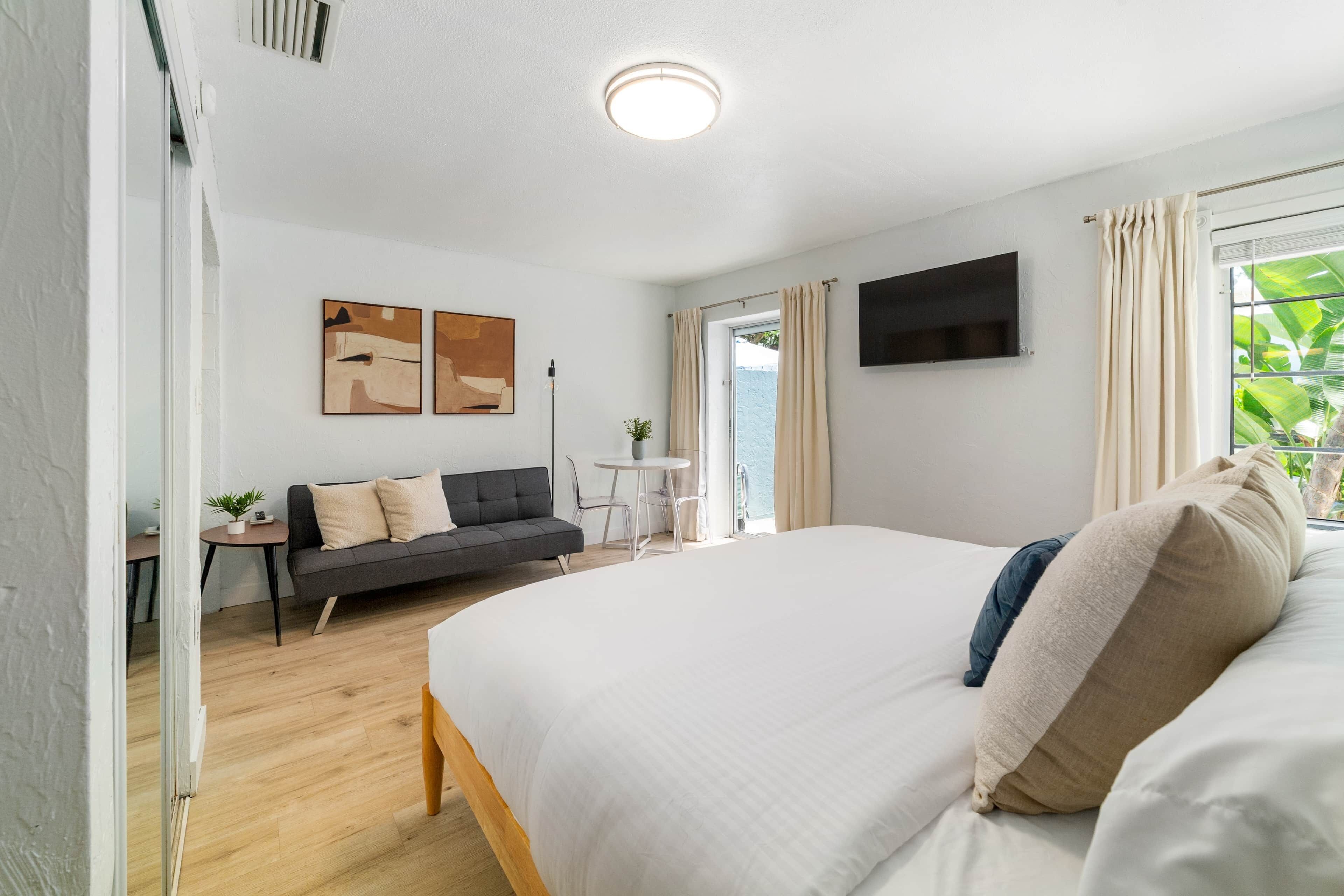A modern bedroom with a large bed, a sofa, a TV mounted on the wall, and a window with curtains.