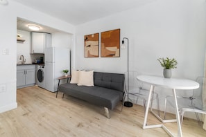 Een moderne keuken met witte kasten, een grijze bank, een ronde tafel met transparante poten en twee abstracte schilderijen aan de muur.
