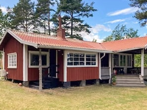 Une cabane en bois rouge avec un porche, une cheminée et un petit jardin devant.