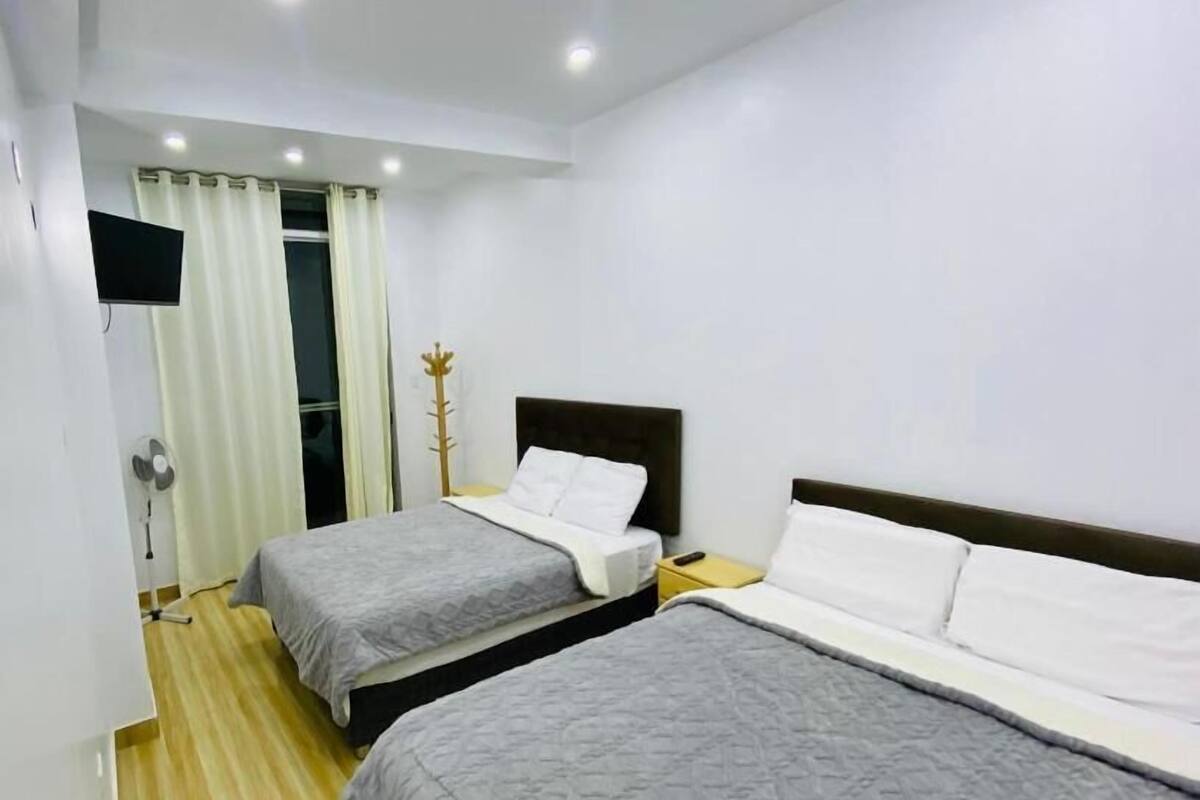 Habitación de hotel con dos camas, un televisor de pantalla plana montado en la pared, piso de madera y una ventana con cortinas.