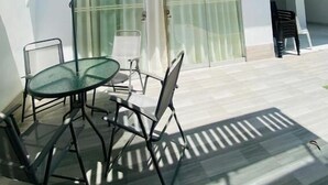 Un área de estar al aire libre moderna con una mesa y sillas de vidrio, un paraguas a rayas y la entrada de un edificio con puerta y cortinas de vidrio.