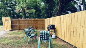 Un jardin avec une clôture en bois, deux chaises à rayures, une table à carreaux et un barbecue.