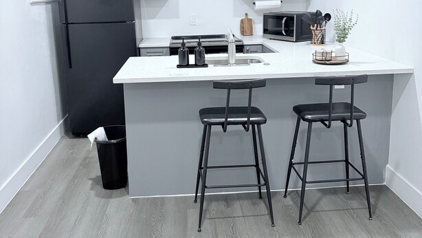 Une cuisine moderne avec des meubles gris, un réfrigérateur noir, un plan de travail blanc et deux tabourets de bar noirs.