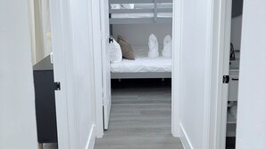 Un couloir étroit mène à une pièce où se trouve un lit superposé, les murs étant peints en blanc et le sol en bois.