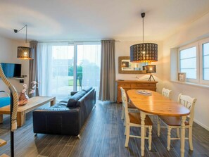 Ein modernes Wohnzimmer mit Sofa, Esstisch, Stühlen, Fernseher und einem großen Fenster mit Vorhängen.