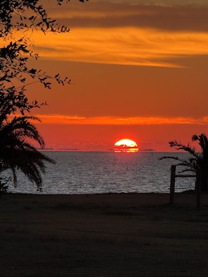 A sunset over a body of water with silhouettes of palm trees and a wooden structure.