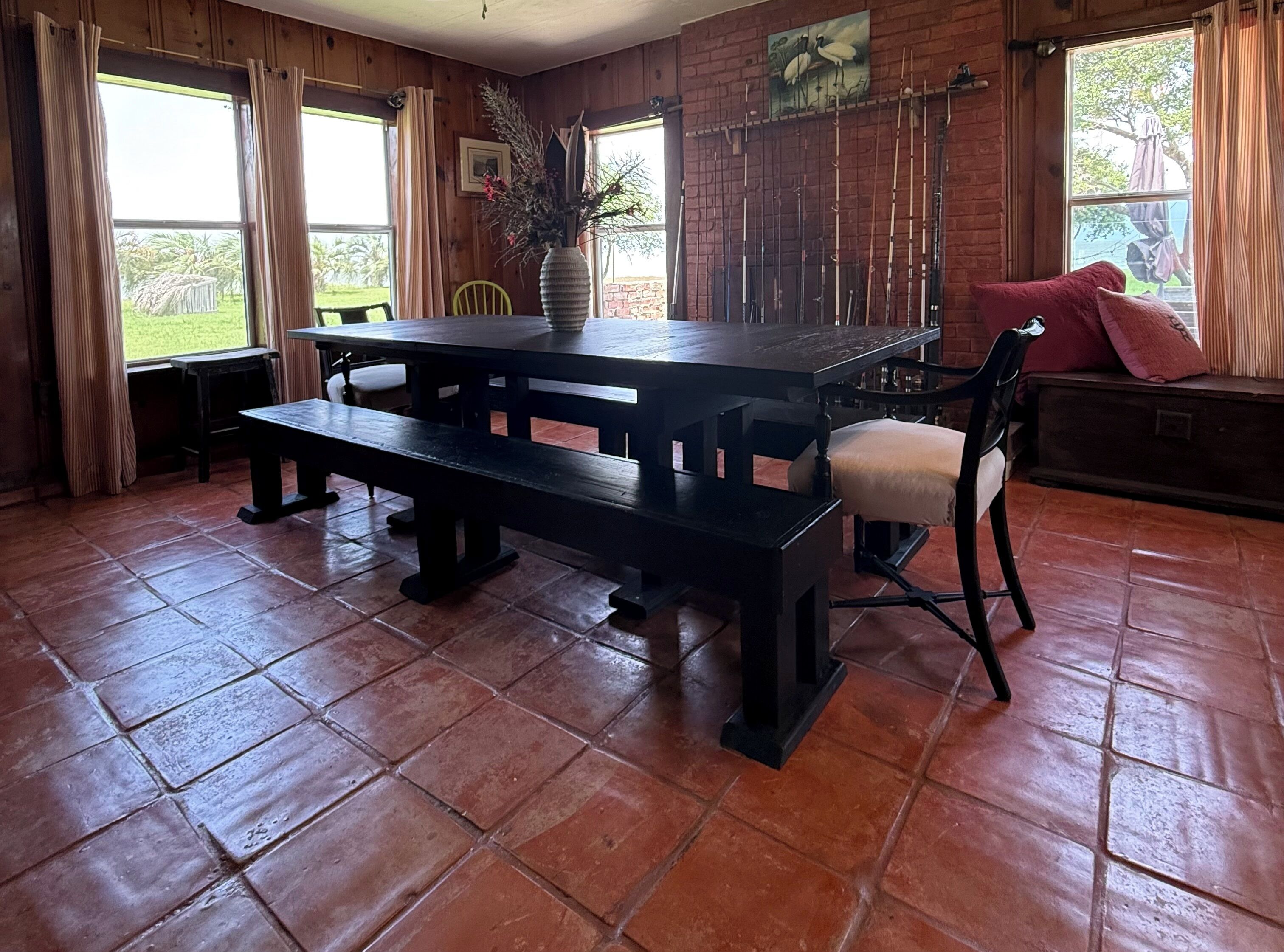Un comedor de madera con una mesa grande y bancos, una silla con cojín y un jarrón con plantas secas.