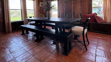 Un comedor de madera con una mesa grande y bancos, una silla con cojín y un jarrón con plantas secas.
