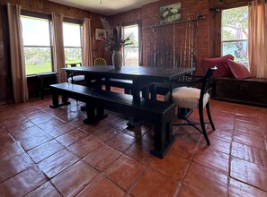 A wooden dining area with a large table and benches, a chair with a cushion, and a vase with dried plants.