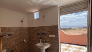 A bathroom with a shower, toilet, and sink, a small window, and a view of a landscape through an open door.