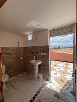 Une salle de bain avec douche, toilettes et lavabo, une petite fenêtre et une vue sur un paysage par une porte ouverte.