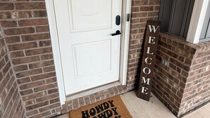 Een witte deur met een keypad, een bruine deurmat met de tekst 'Howdy Howdy', en een houten bord waarop 'WELCOME' staat.