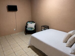 A bedroom with a bed, a chair, a small table, a TV mounted on the wall, and a tiled floor.