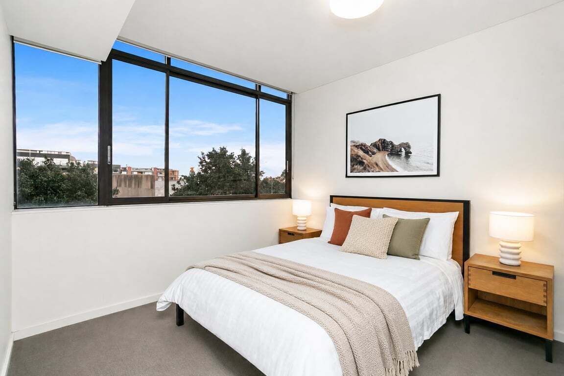 Een slaapkamer met een groot raam, een bed met houten hoofdeinde, twee nachtkastjes met lampen, en een lijst met een afbeelding aan de muur.