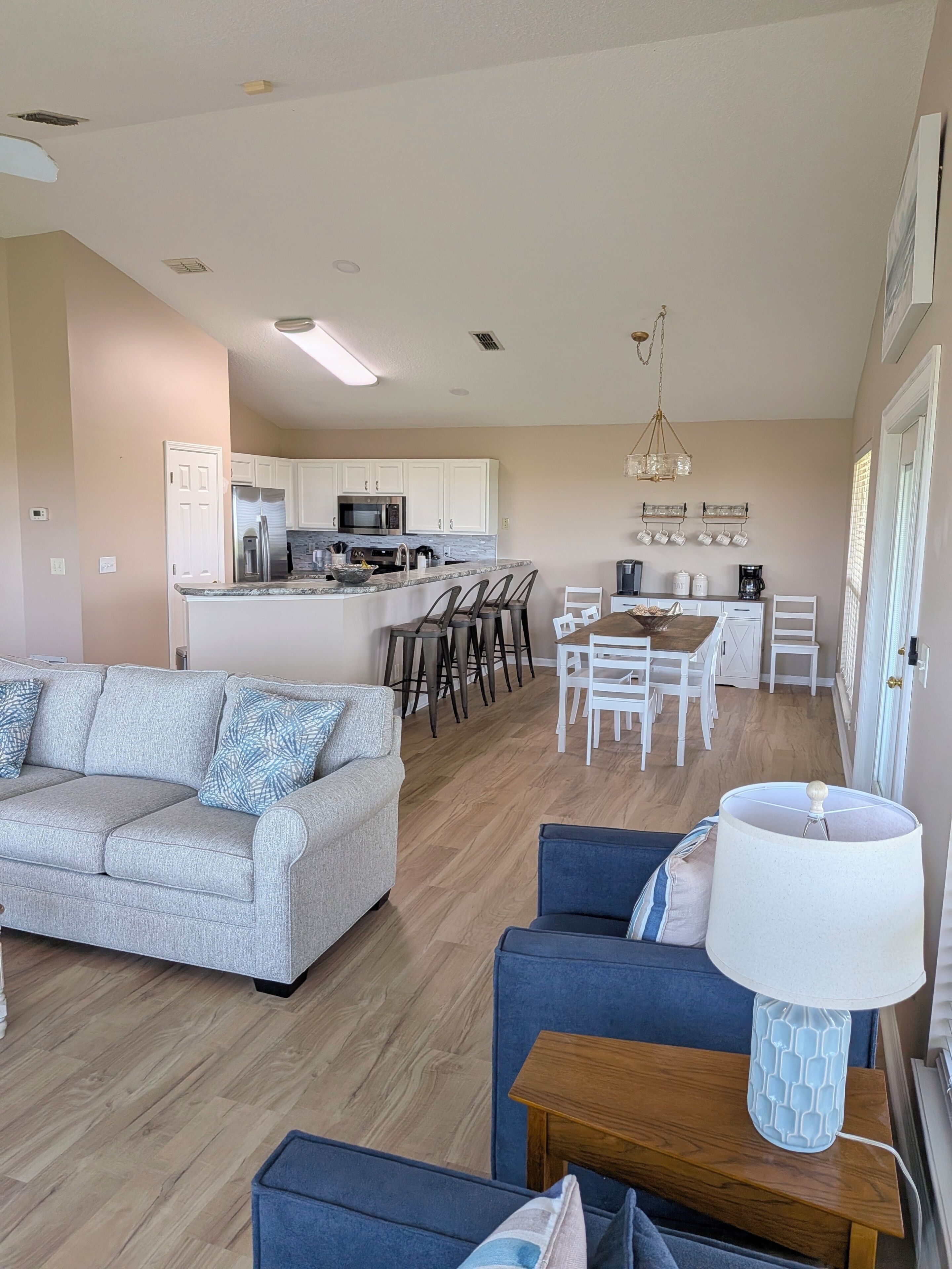 A modern living room with a grey sofa, blue armchairs, and a wooden coffee table. A kitchen with white cabinets and a dining area with white chairs is visible in the background.