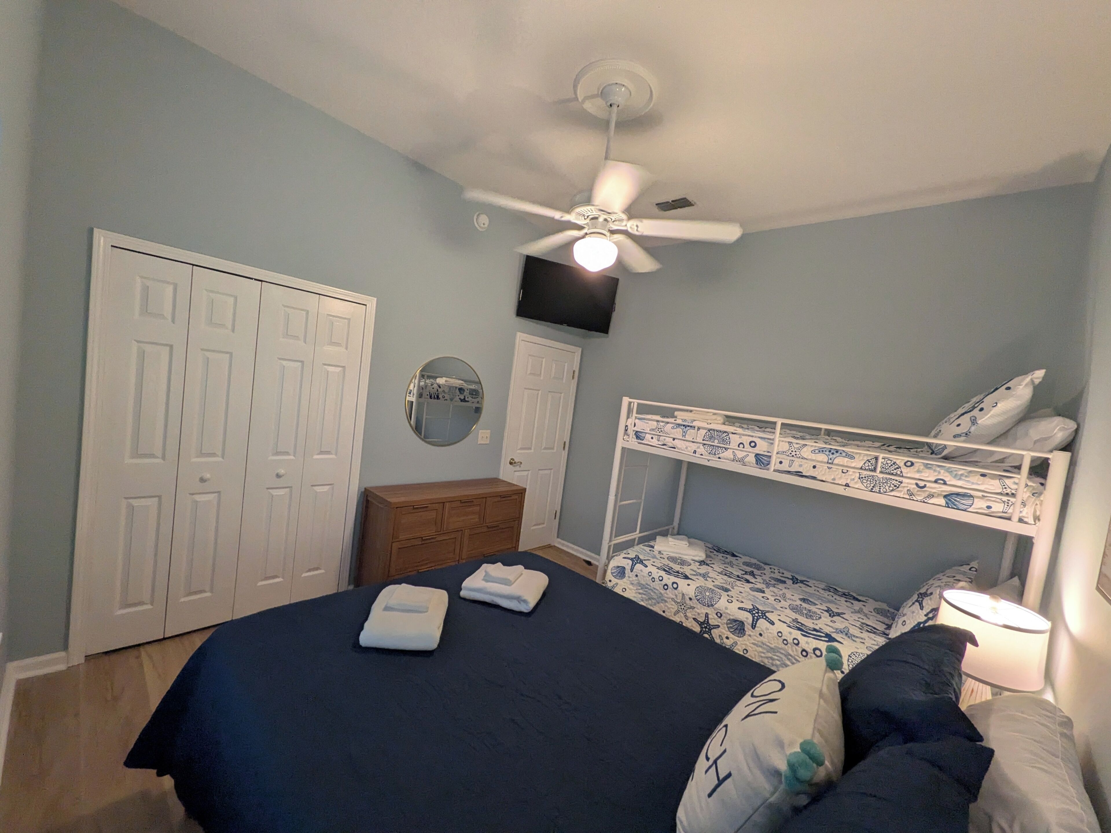 Un dormitorio con una cama litera, ventilador de techo, cómoda y espejo redondo.