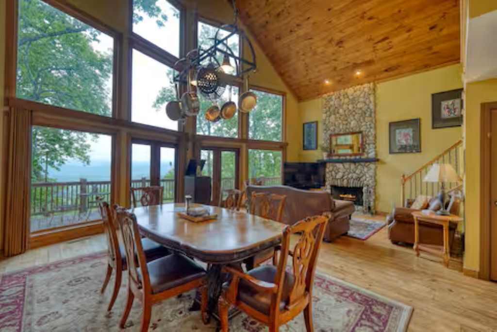 Un comedor amplio con una gran mesa y sillas de madera, una chimenea de piedra y vistas a través de amplios ventanales.