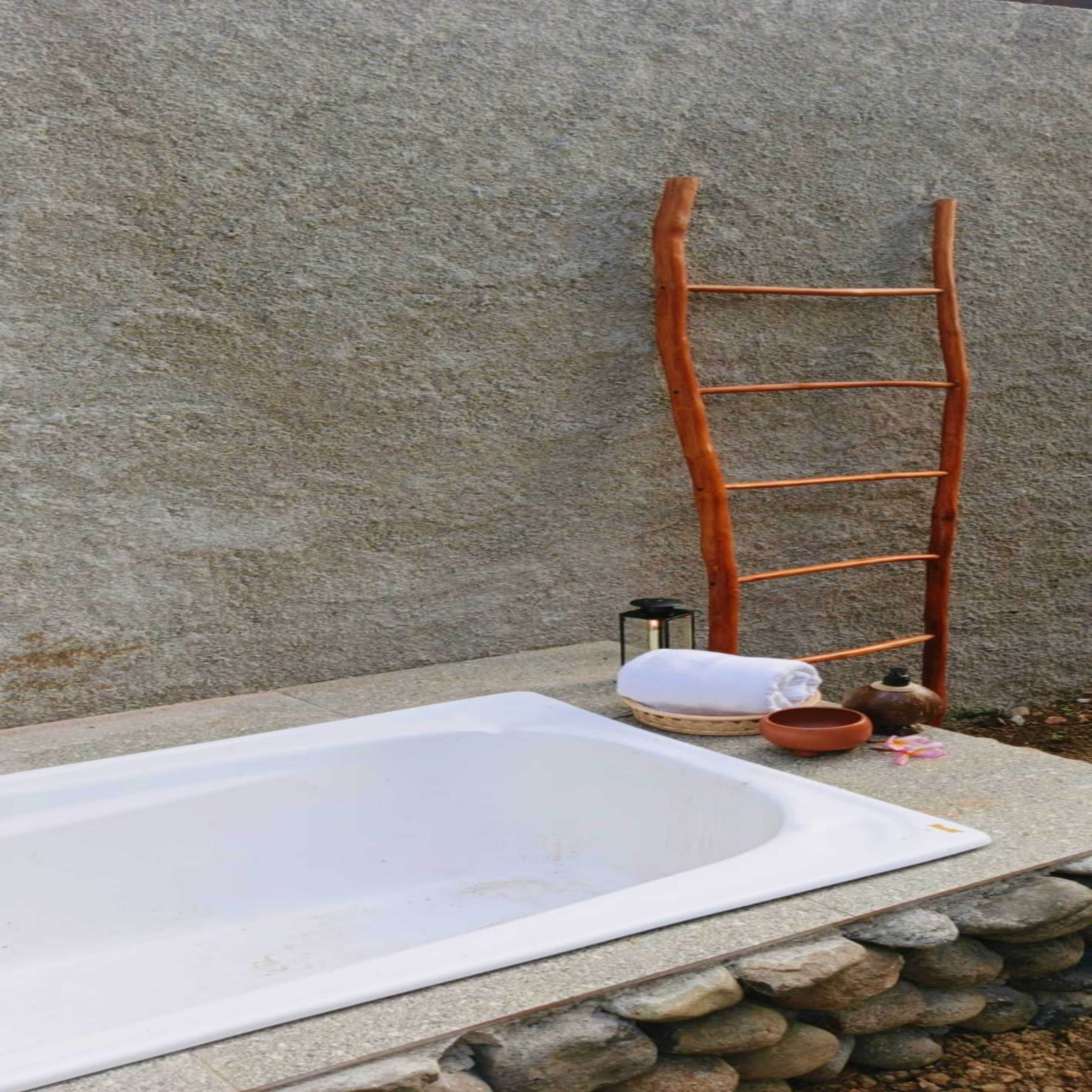 A stone wall, a wooden ladder, a white bathtub, a white towel, a small lantern, and a clay pot.