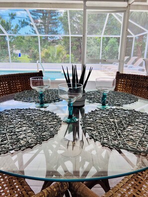 A round glass table with woven placemats, two glass vases with black decorative items, and a view of a pool and lounge chairs through large windows. - Spacious Florida Pool Retreat - 15 min from beach (Vero Beach)