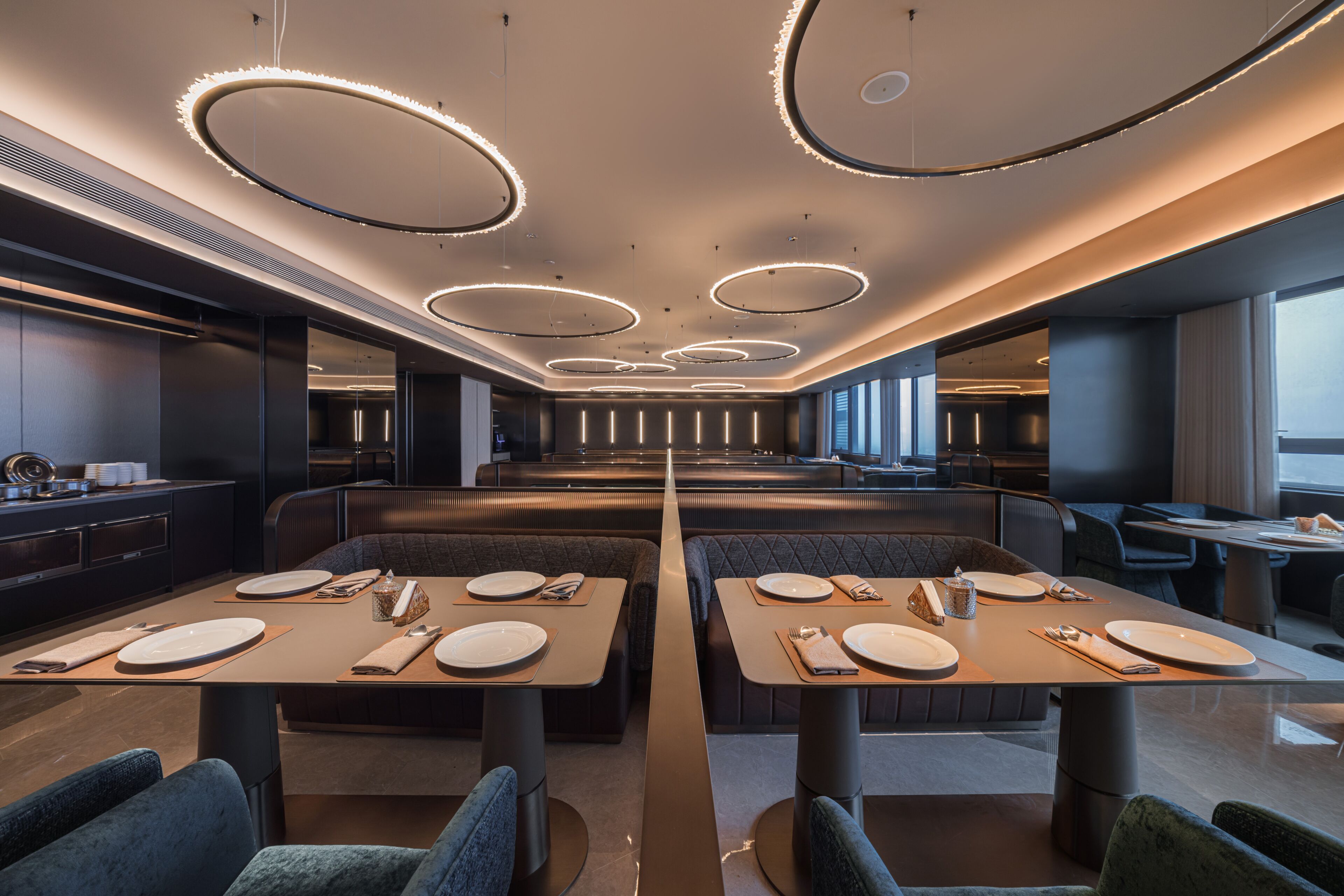 A modern dining area with circular ceiling lights, long tables, and cushioned booths.
