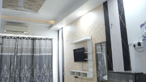A room with a bed, a flat-screen TV mounted on the wall, a refrigerator, and an air conditioning unit. - Arista Stays (Greater Noida)