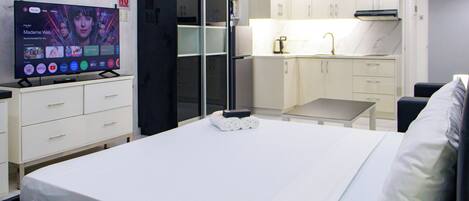 Habitación de hotel moderna con cama, televisor mostrando un servicio de streaming, kitchenette y zona de estar.