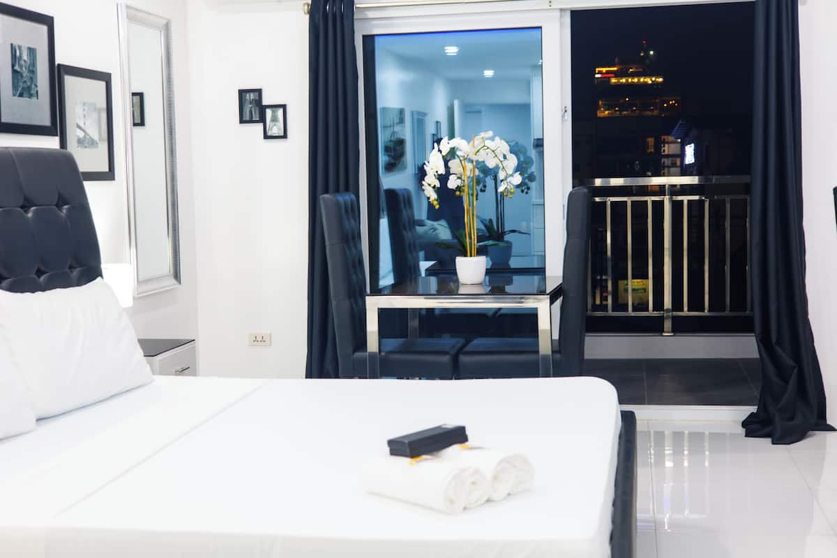 A hotel room with a bed, a desk with a potted orchid, a chair, a window with curtains, and a balcony with a railing.