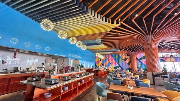 A modern restaurant with a vibrant interior, featuring a central island with seating, a colorful geometric wall design, and a unique ceiling structure. - Flamingo Hai Tien Beach Resort (Hoang Tien)