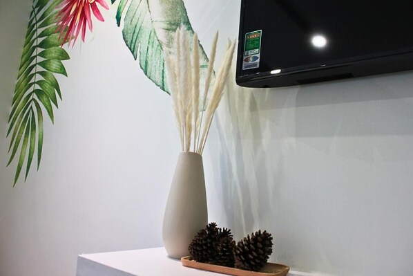 Un téléviseur fixé au mur, un vase blanc contenant des plantes séchées et un plateau en bois avec des pommes de pin, le tout posé sur une surface blanche.