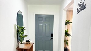 Un couloir avec un banc en bois, un coussin décoratif sur le thème de l’accueil, une plante en pot, un miroir et une porte avec un paillasson.